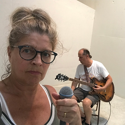 Photo of woman holding a microphone and looking at the camera, with a man sitting down and holding an electric guitar in the background (Lilly Santon and Jimmy Weinstein)