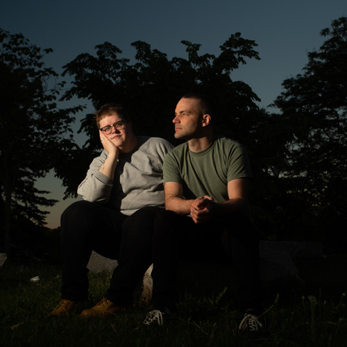 Photo of two men sitting outside, one looking at the camera and the other looking away (Joyful Joyful)